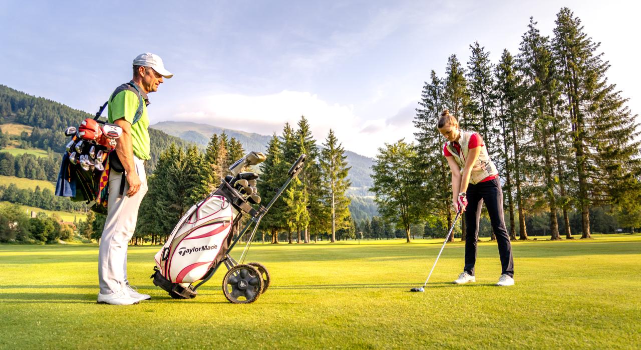 Ein Golfer beobachtet eine Frau, die sich auf einen Golfschlag vorbereitet, umgeben von Bergen