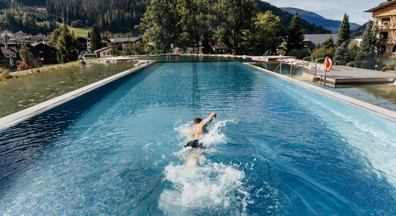 A swimmer takes a dip in an impressive outdoor pool surrounded by a picturesque mountain landscape. The lush trees and rolling hills provide a tranquil backdrop, while the clear water reflects the light of the sun.