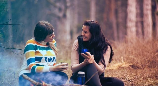126cf3e0-544f-4678-b41f-ff82d484e32f Zwei Frauen sitzen im Wald vor einem Lagerfeuer und trinken aus einer Thermoskanne
