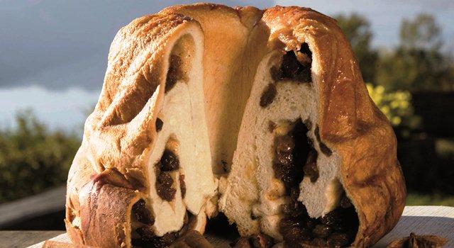 Kärntner Reindling mit Trockenfrüchten Ein traditioneller Kärntner Reindling, angeschnitten, zeigt seine reichhaltige Füllung aus getrockneten Früchten. Der Guglhupf ist goldbraun gebacken und umgeben von einer idyllischen Landschaft, die dem Bild eine rustikale und gemütliche Atmosphäre verleiht.