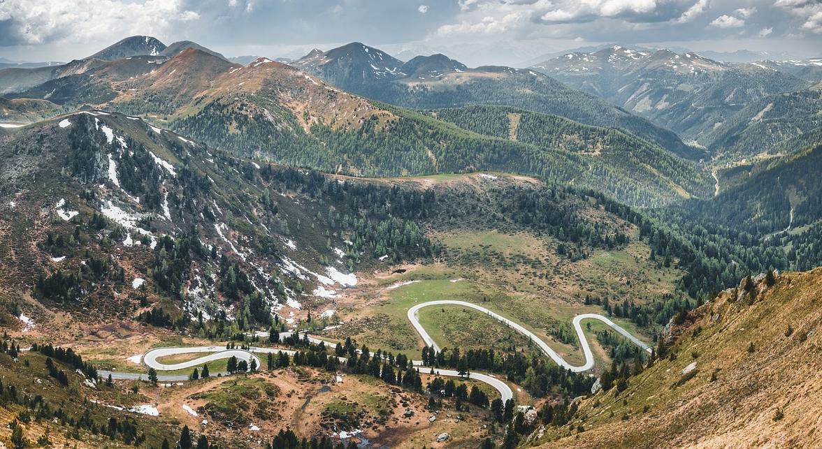 915fc958-7fab-4c23-90d8-9a0544b94d97 Eine kurvenreiche Bergstraße schlängelt sich durch eine grüne und teilweise schneebedeckte Berglandschaft