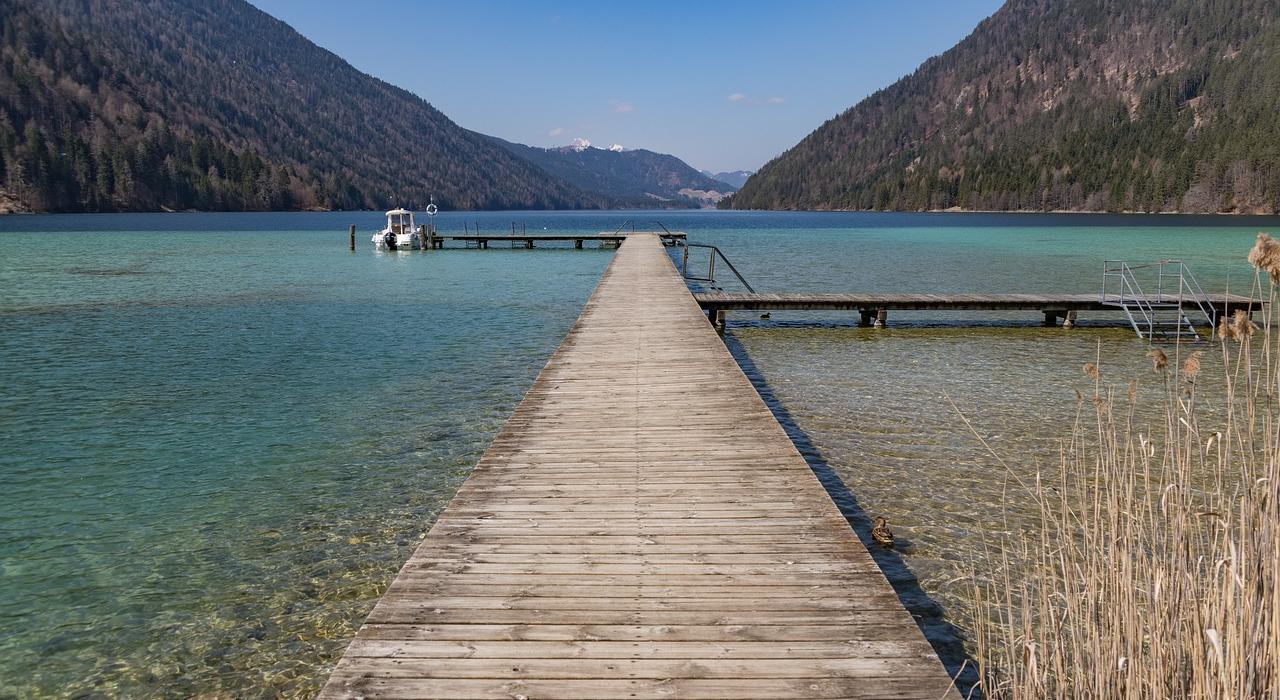 06882b53-de9e-427c-a810-ef5887c7bc27 Ein Holzsteg, der in einen klaren Bergsee führt, umgeben von bewaldeten Hügeln und Bergen im Hintergrund. Ein Boot ist am Ende des Stegs festgemacht