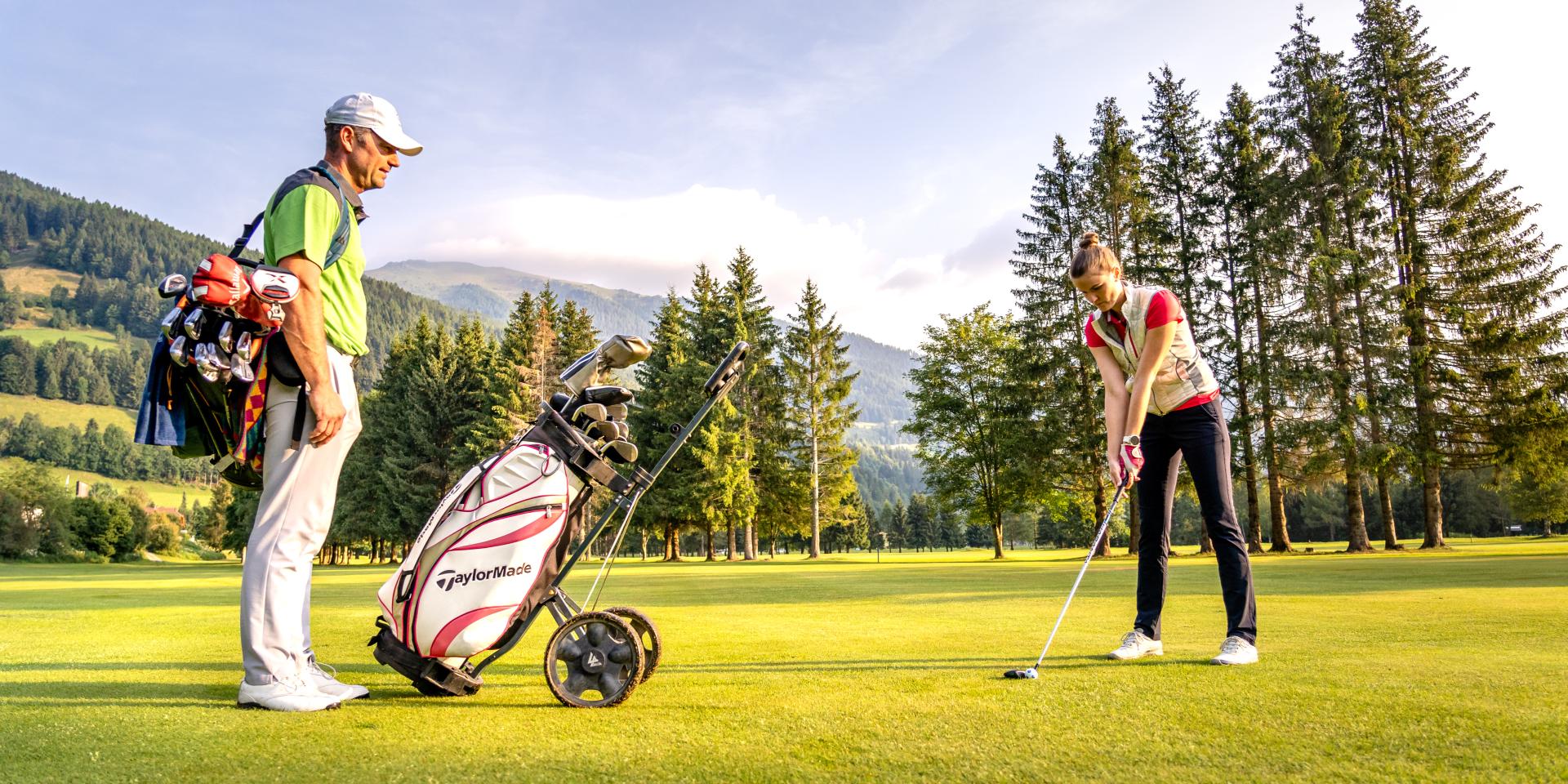 Golf_Bad Kleinkirchheim_Sommer © Mathias Prägant_MBN Tourismus(52) Ein Golfer beobachtet eine Frau, die sich auf einen Golfschlag vorbereitet, umgeben von Bergen