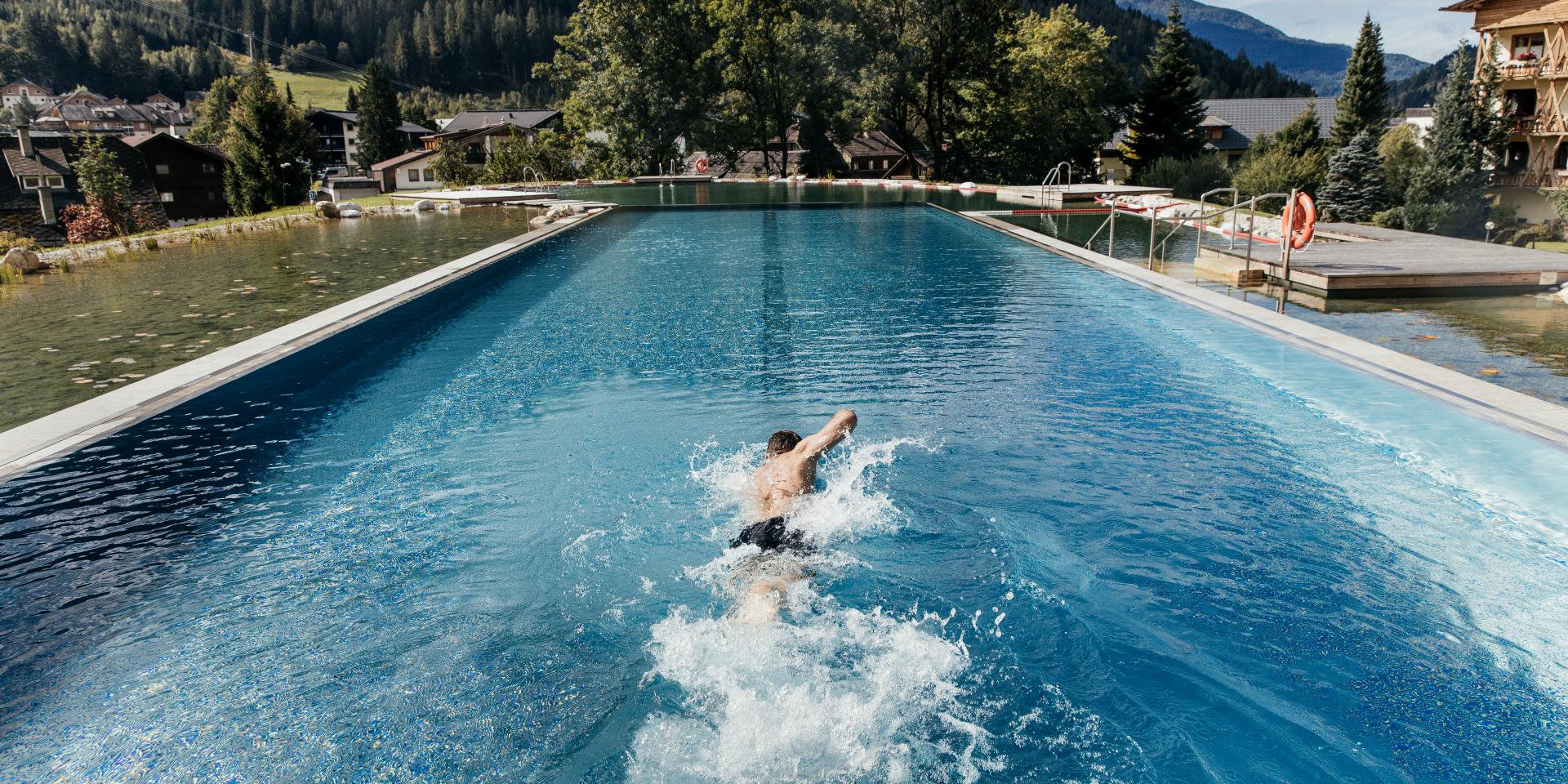 Ein Schwimmer zieht seine Bahnen in einem beeindruckenden Außenpool, umgeben von einer malerischen Berglandschaft. Die üppigen Bäume und sanften Hügel bieten eine ruhige Kulisse, während das klare Wasser das Licht der Sonne reflektiert.