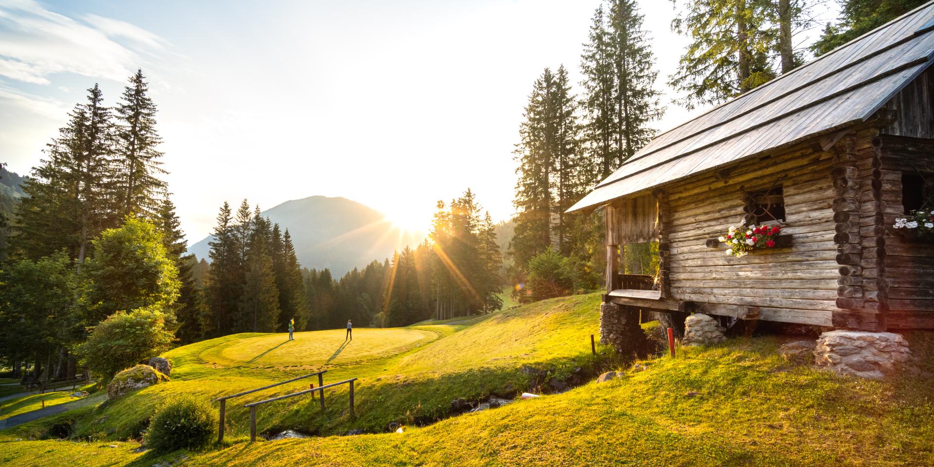 Golf_Bad Kleinkirchheim_Sommer © Mathias Prägant_MBN Tourismus(2) Ein Holzhaus bei Sonnenuntergang und in der Ferne Menschen beim Golfspielen im Sommerurlaub in Kärnten