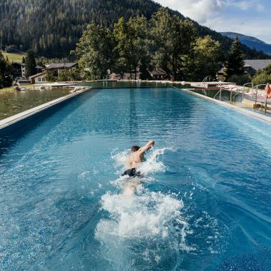 A swimmer takes a dip in an impressive outdoor pool surrounded by a picturesque mountain landscape. The lush trees and rolling hills provide a tranquil backdrop, while the clear water reflects the light of the sun.