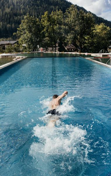 Ein Schwimmer zieht seine Bahnen in einem beeindruckenden Außenpool, umgeben von einer malerischen Berglandschaft. Die üppigen Bäume und sanften Hügel bieten eine ruhige Kulisse, während das klare Wasser das Licht der Sonne reflektiert.