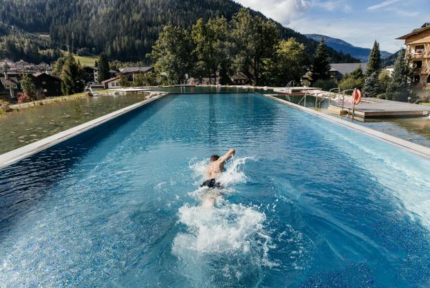 A swimmer takes a dip in an impressive outdoor pool surrounded by a picturesque mountain landscape. The lush trees and rolling hills provide a tranquil backdrop, while the clear water reflects the light of the sun.