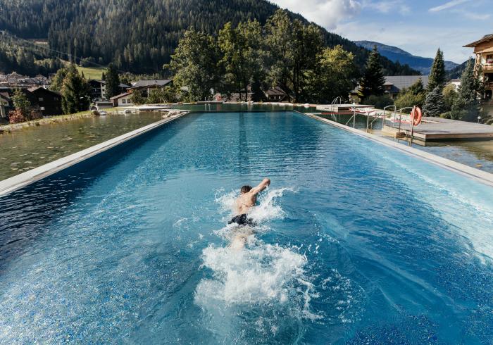 Ein Schwimmer zieht seine Bahnen in einem beeindruckenden Außenpool, umgeben von einer malerischen Berglandschaft. Die üppigen Bäume und sanften Hügel bieten eine ruhige Kulisse, während das klare Wasser das Licht der Sonne reflektiert.